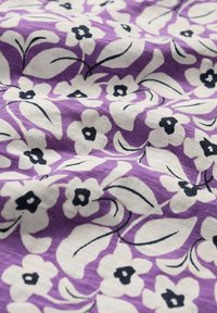 Tela morada con un patrón floral que presenta flores blancas y centros negros. Las hojas están contorneadas en negro, creando un contraste en la textura suave.