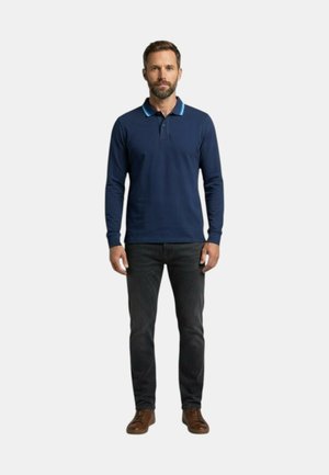 Uomo in piedi rivolto verso lo spettatore, indossa una polo blu scuro a maniche lunghe, jeans scuri e scarpe marroni su uno sfondo semplice.