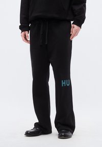 Μαύρο παντελόνι φόρμας με άνετη εφαρμογή, με ελαστική μέση και κορδόνια, διακοσμημένο με μια τιρκουάζ γραφική παράσταση "HU" στο αριστερό πόδι.