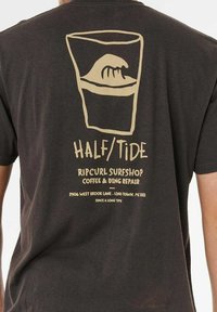 Donkergrijze katoenen T-shirt met een beige grafiek van een golf in een glas, met de tekst "HALF/TIDE" en "RIPCURL SURFSHOP" eronder. Eenvoudig ontwerp.