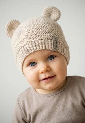Kleinkind mit blauen Augen, das eine beige gestrickte Mütze mit Bommelohren und ein hellbraunes Shirt trägt, lächelt sanft vor einem schlichten Hintergrund.