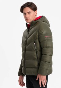 Olivgrön puffjacka med huva, med chevron-quiltning, två dragkedjefickor på sidorna och ett rött innerfoder.