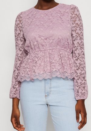 Person trägt eine langärmelige lilafarbene Spitzenbluse mit Blumenmuster und hellblaue hoch taillierte Jeans, dargestellt vom Hals bis zur Hüfte.
