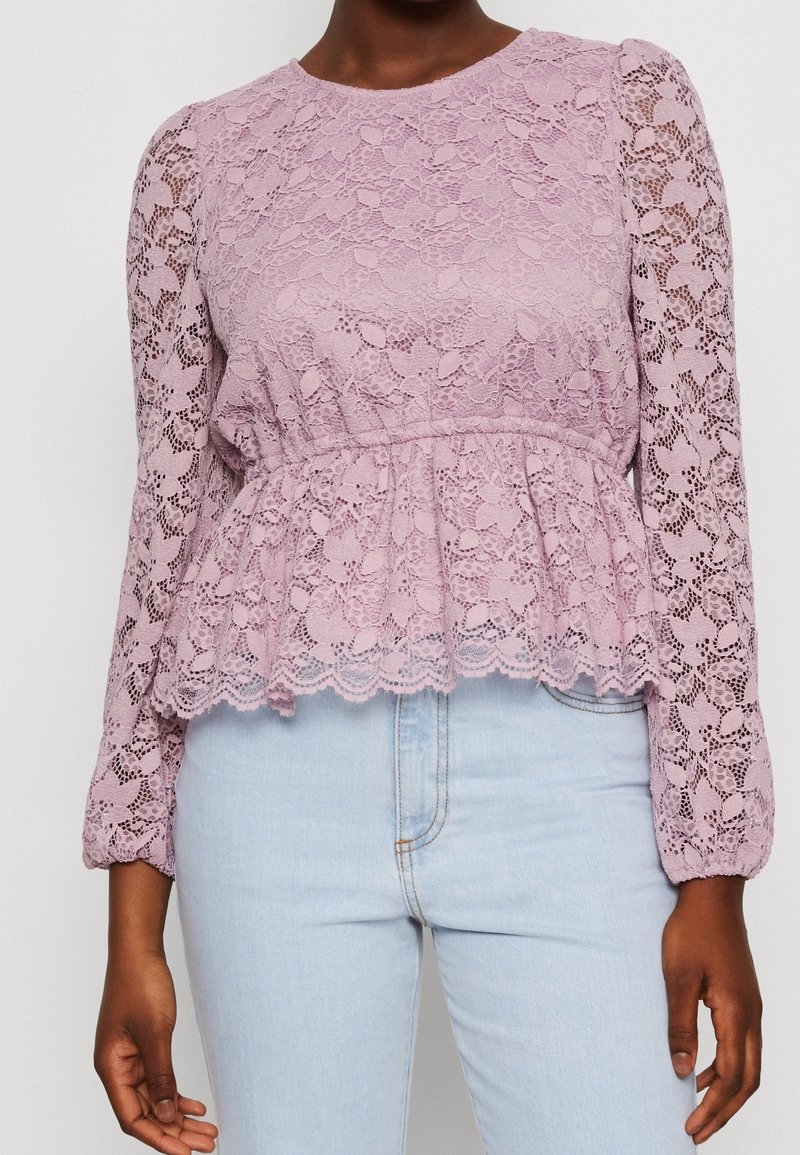 Personne portant un chemisier en dentelle lilas à manches longues avec un motif floral et un jean bleu clair taille haute, montré du cou aux hanches.
