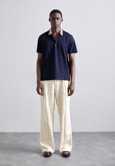 Polo bleu marine avec col rayé multicolore, associé à un pantalon ample crème et des chaussures marron foncé. Design minimaliste et coupe classique.