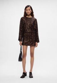 Smeđa haljina sa šljokicama, dugih rukava, kroja koji se sužava u struku, dužine iznad koljena. Uparena s crnom torbom i sjajnim cipelama na petu.