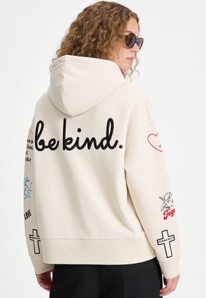 Femme aux cheveux bouclés portant des lunettes de soleil et un sweat à capuche crème avec le texte "be kind." et divers petits graphismes dans le dos et sur les manches.