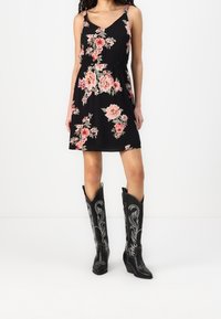 Woman wearing a sleeveless black dress with pink floral print and black knee-high western boots with white embroidery.