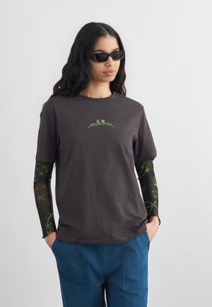 Γυναίκα με μακριά μαλλιά που φορεί μαύρα γυαλιά ηλίου, μπλούζα γραφικών σκούρου γκρι χρώματος με μοτίβα στα μανίκια δίχτυ, και μπλε παντελόνι, με τα χέρια στις τσέπες.