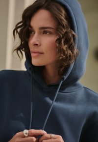 Dunkelblauer Hoodie mit Kordelzug, weicher Textur und lockerer Passform. Auffällige Details sind eine große Kapuze und mehrfach geschichtete Ringe an den Fingern.