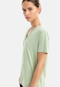 Jasnozielona koszulka z krótkim rękawem, z dekoltem w serek, o miękkiej teksturze materiału i luźnym kroju. Brak widocznych wzorów lub akcentów.