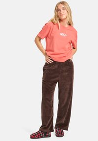 Koraalkleurig T-shirt met een wit logo, bruine corduroy broek en rode geruite pantoffels. De broek heeft een ontspannen pasvorm en een elastische tailleband.