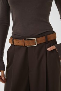 Cintura in pelle scamosciata marrone con fibbia in metallo argento, dotata di fori regolabili. Indossata sopra pantaloni di tessuto marrone scuro con una maglia fitted a maniche lunghe.