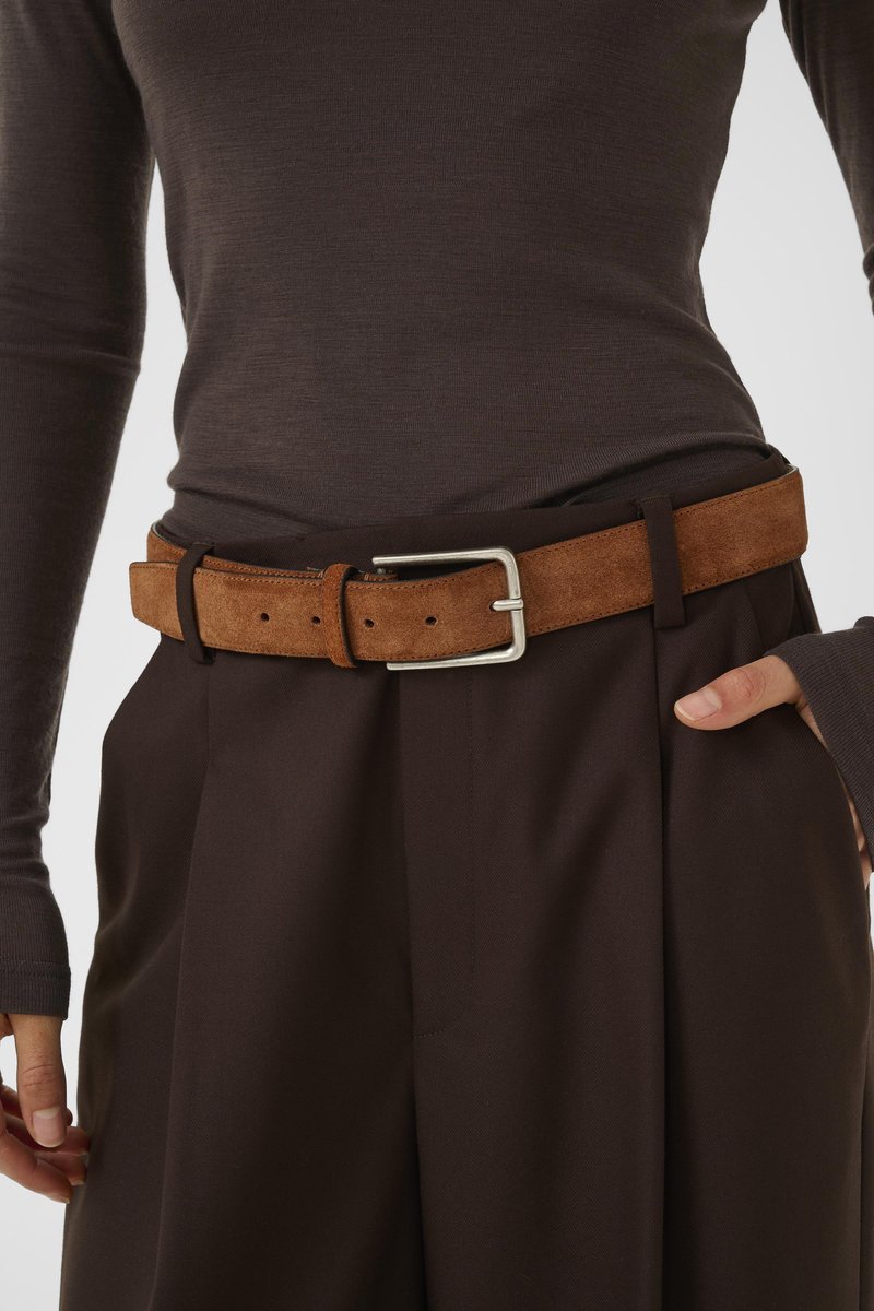 Cintura in pelle scamosciata marrone con fibbia in metallo argento, dotata di fori regolabili. Indossata sopra pantaloni di tessuto marrone scuro con una maglia fitted a maniche lunghe.