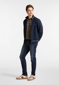 Chaqueta de punto navy con tejido texturizado y acentos marrones; camiseta oliva con texto impreso; jeans azul oscuro; zapatillas beige y marrones.