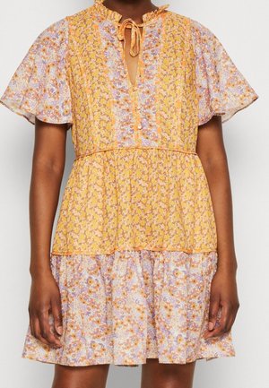 Femme portant une robe courte à manches courtes, à motifs floraux jaunes et violets, avec un col à volants et un nœud à l'encolure.