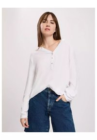 Biała koszula z długim rękawem, z dekoltem w serek i trzema guzikami, w połączeniu z szortami dżinsowymi z wysokim stanem. Miękka tkanina o luźnym kroju.
