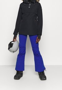 Czarna kurtka z kapturem i futrzanym obszyciem, zestawiona z niebieskimi spodniami narciarskimi i czarnymi butami. W lewej ręce trzymany jest szary kask.