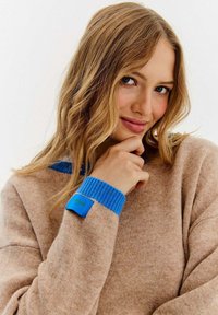 Beżowy sweter o fakturowanej powierzchni, z jasnoniebieskim ściagaczem przy kołnierzu i mankietach oraz niebieską metką z zielonym tekstem na rękawie.