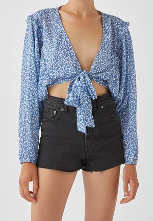 Blouse courte bleu clair à motif floral, fermeture nouée à l'avant, manches longues et tissu texturé ; associée à un short en denim noir.