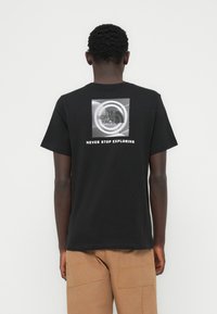 Μαύρο βαμβακερό μπλουζάκι με γραφικό σχέδιο στην πλάτη, που περιλαμβάνει κυκλικά μοτίβα και το κείμενο «ΠΟΤΕ ΜΗΝ ΣΤΑΜΑΤΑΣ ΝΑ ΕΞΕΡΕΥΝΑΣ».