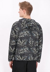 Hooded jack met een zwarte basis en een groen-blauw bladerenpatroon. Gemaakt van lichtgewicht stof met een gladde textuur en een relaxte pasvorm.