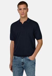 Jeune homme portant un polo bleu marine à manches courtes et un jean bleu clair, debout avec une main dans la poche sur un fond uni.