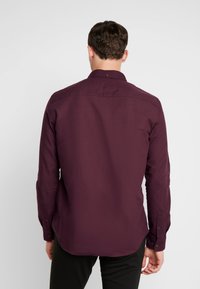 Man met de rug naar de camera, gekleed in een donkerpaarse overhemd met lange mouwen en zwarte broek, tegen een effen witte achtergrond.