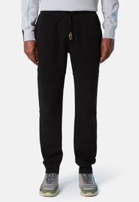Pantalones de chándal negros con cintura ajustable, que presentan una textura suave y bolsillos laterales, combinados con zapatillas en tonos gris y verde.