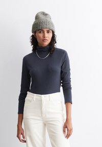 Tumed turtleneck pluus pika varrukaga, kantud kreemivärvi korgitud pükstega. Mudel kannab kudumit roheline beanie ja hõbedast kaelakeed.