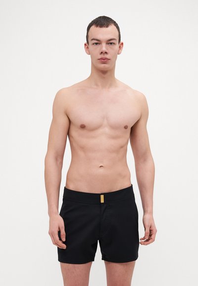 Shorts de bain noirs avec une texture lisse, ornés d'un accent doré à l'avant et d'une taille élastique. Le modèle se tient torse nu contre un fond uni.