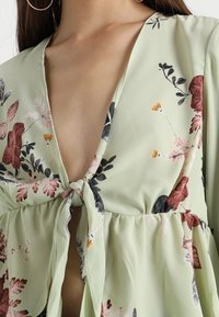 Lysegrøn blomstret bluse med dyb V-udskæring, båndknude detalje foran, med multifarvede blomsterprint og lette ærmer.