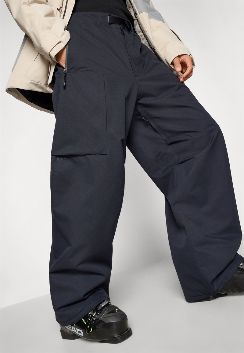 Navy cargo pants with a relaxed fit, featuring large side pockets, smooth fabric, and adjustable waistband. Worn with black ski boots.