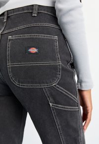 Pantalones de mezclilla negros con costuras en contraste, con un bolsillo trasero que tiene una etiqueta de logo. Diseño de corte recto y múltiples bolsillos funcionales.