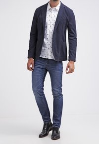 Marinblazer över en vit skjorta med svarta prickar, ihop med blå jeans och svarta läderskor. Har en skräddarsydd passform och en avslappnad stil.