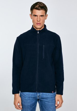 Jauns vīrietis, kas valkā tumši zilu zip-up flīsa jaku un zilas džinsas, stāv pret vienkāršu gaišu fonu.