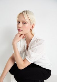 Valge pluus puffed sleeves'iga, mustad püksid. Mudel poseerib käsi lõua all, sile nahk, sirged blondid juuksed, neutraalne taust.