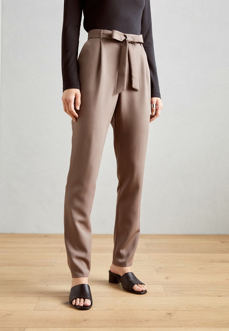 Pantalons marron taille haute avec un nœud à la taille, coupe ajustée, tissu lisse, associés à des sandales à lanières noires ouvertes.