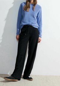 Vaaleansininen neulepaita, mustat leveälahkeiset housut, teksturoitu kangas, kiristysnauhavyötärö, yhdistetty mustiin lenkkareihin. Seisoo valkoista seinää vasten.