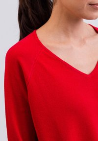 Roter, gestrickter Pullover mit V-Ausschnitt, langen Ärmeln und einer weichen Textur. Der Stoff wirkt warm und leicht dehnbar.
