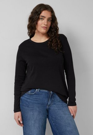 Junge Frau mit langen lockigen Haaren, die ein schwarzes Langarmshirt und blaue Jeans vor einem einfarbigen grauen Hintergrund trägt.