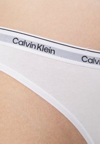 Hvit bomullsthong med en flat midjebånd med en svart og grå "Calvin Klein" logo. Glatt tekstur med minimale sømmer.