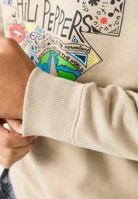 Beige sweater med grafisk design, der fremviser farverige verdenskort og tekstaccent. Ribbed ærmer og blød stofstruktur.