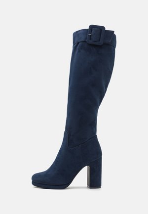 Bota de ante azul marino hasta la rodilla con tacón cuadrado, que presenta un puño ancho y un detalle de hebilla cuadrada en el lado exterior. Textura suave.