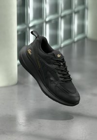 Μαύρο αθλητικό παπούτσι Lacoste με χρυσό λογότυπο και κορδόνια, αιωρούμενο πάνω από λεία επιφάνεια κοντά σε τοίχο από γυάλινα τούβλα σε μοντέρνο εσωτερικό χώρο.