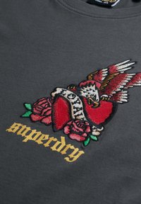 Sudadera gris con un destacado diseño bordado de un corazón, un águila y rosas, con "Superdry" en fuente dorada en la parte inferior.