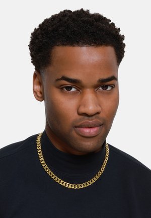 Collier en chaîne dorée avec de gros maillons entrelacés, porté sur une chemise noire à col haut ; met en valeur une finition métallique lisse et un design audacieux.