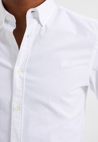 Biała koszula na guziki z usztywnianym kołnierzykiem, lekkim materiałem, subtelnym teksturowanym wykończeniem oraz małym haftowanym logo na piersi.