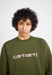 Felpa verde oliva con scollo rotondo, caratterizzata da un logo "carhartt" ricamato in rosa sulla parte anteriore. Tessuto morbido e vestibilità comoda.