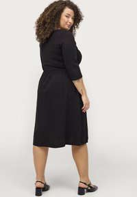 Zwarte jurk tot op de knie met driekwartmouwen, geplooide taille en een zachte textuur, gecombineerd met zwarte gehakte sandalen.